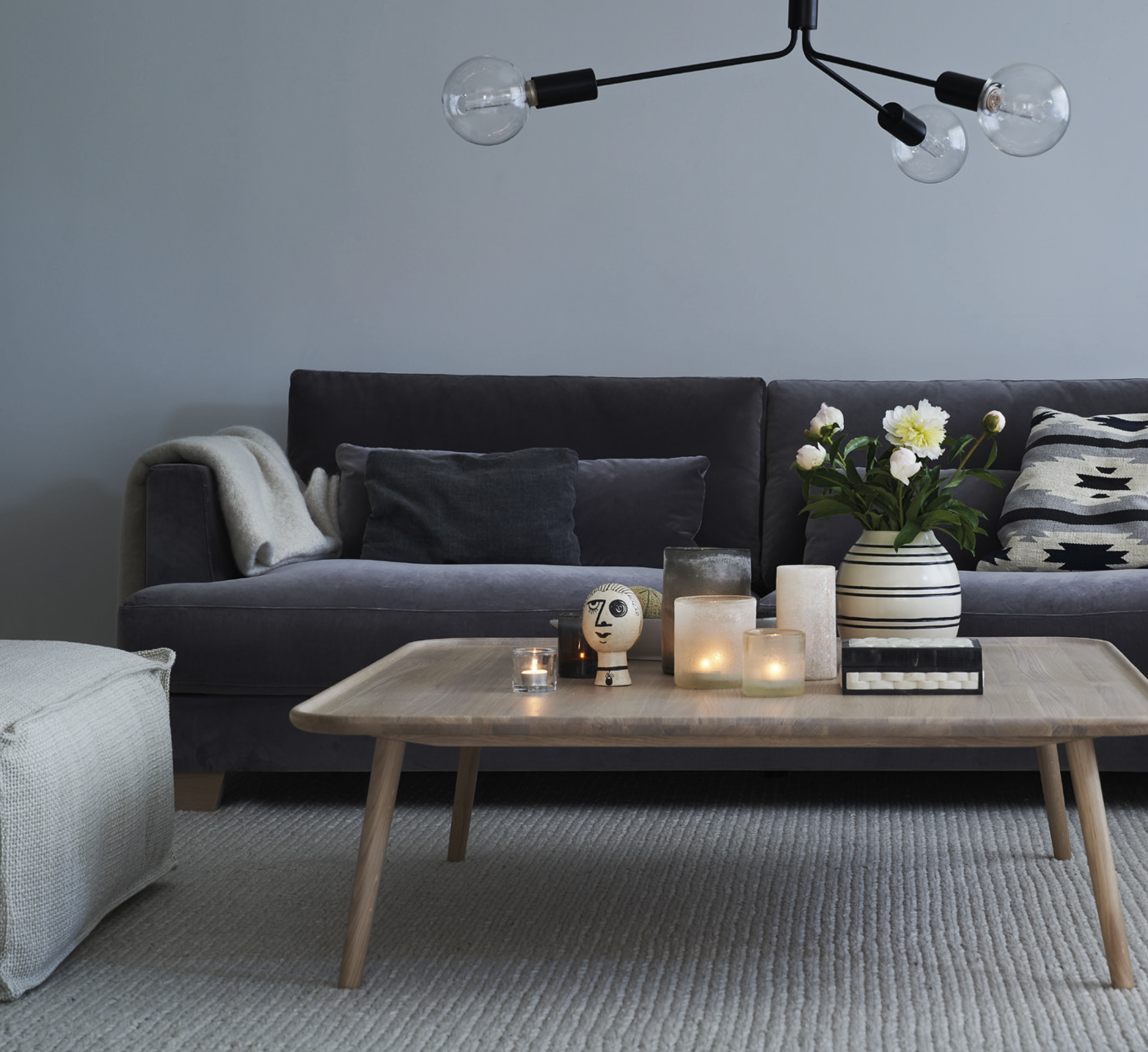 Salon moderne avec un canapé gris, une table basse en bois, des bougies, des fleurs et un plafonnier. Canape Paris by Atelier Bis