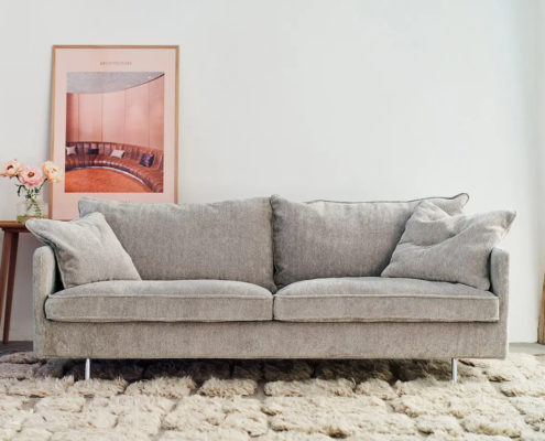 Canapé gris clair sur un tapis texturé avec une table d'appoint, des fleurs et un tableau encadré sur un mur blanc. Canape Paris by Atelier Bis