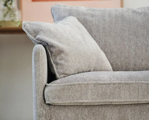 Gros plan sur un canapé gris clair avec un coussin, un tapis en fourrure, des fleurs et des œuvres d'art en arrière-plan. Canape Paris by Atelier Bis