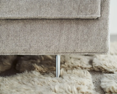 Gros plan sur un canapé beige avec un pied en métal, posé sur un tapis moelleux de couleur claire. Canape Paris by Atelier Bis