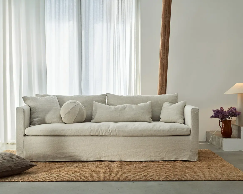 Un canapé beige avec des coussins repose sur un tapis tressé dans un salon lumineux et minimaliste avec des rideaux transparents. Canape Paris by Atelier Bis