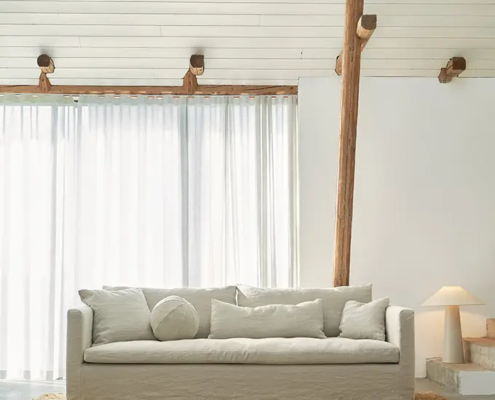 Salon minimaliste avec un canapé blanc, un coussin rond, un tapis de jute et une lumière naturelle douce grâce à des rideaux transparents. Canape Paris by Atelier Bis