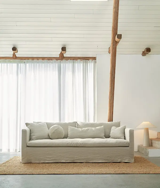 Salon minimaliste avec un canapé blanc, un coussin rond, un tapis de jute et une lumière naturelle douce grâce à des rideaux transparents. Canape Paris by Atelier Bis