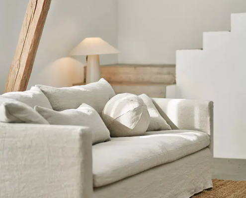 Un salon minimaliste avec un canapé blanc, des coussins neutres, une lampe de table et des touches de bois naturel. Canape Paris by Atelier Bis