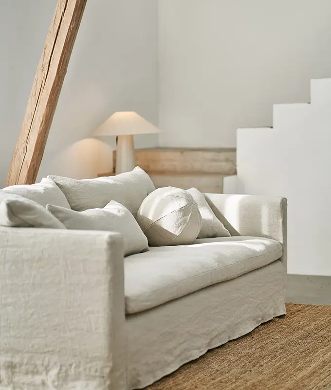 Un salon minimaliste avec un canapé blanc, des coussins neutres, une lampe de table et des touches de bois naturel. Canape Paris by Atelier Bis