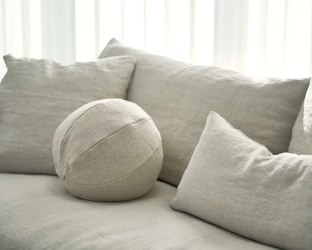 Canapé beige avec des coussins carrés et ronds assortis devant des rideaux blancs transparents. Canape Paris by Atelier Bis