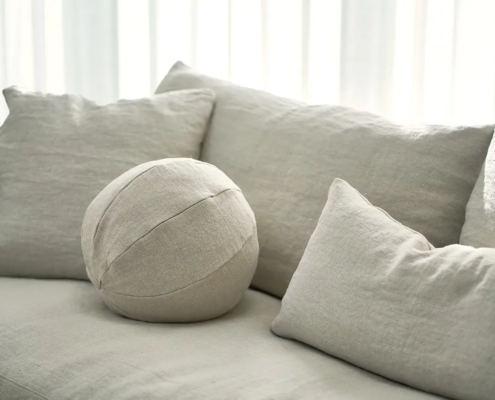 Canapé beige avec des coussins carrés et ronds assortis devant des rideaux blancs transparents. Canape Paris by Atelier Bis