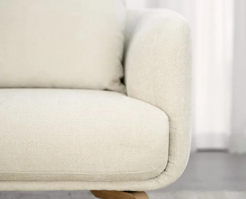Gros plan sur un canapé moderne beige avec des pieds en bois, posé sur un tapis de couleur claire, devant des rideaux transparents. Canape Paris by Atelier Bis
