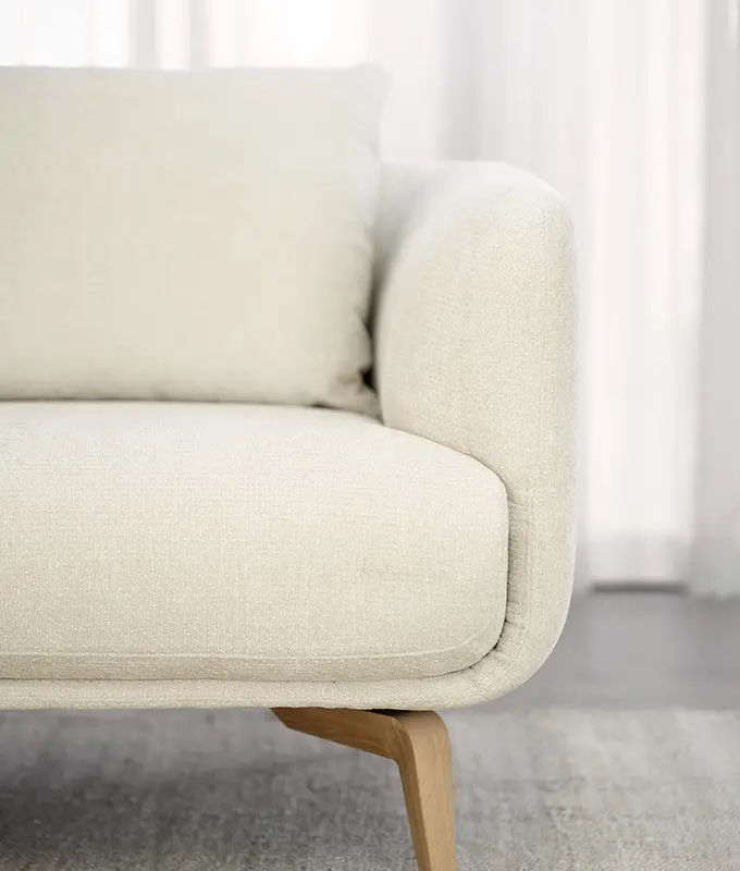 Gros plan sur un canapé moderne beige avec des pieds en bois, posé sur un tapis de couleur claire, devant des rideaux transparents. Canape Paris by Atelier Bis