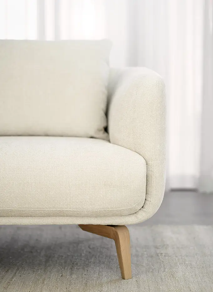 Gros plan sur un canapé moderne beige avec des pieds en bois, posé sur un tapis de couleur claire, devant des rideaux transparents. Canape Paris by Atelier Bis