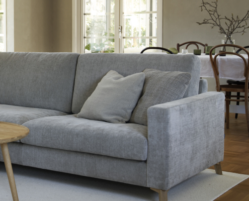 Canapé gris clair avec deux oreillers dans un salon, table basse en bois et salle à manger en arrière-plan. Canape Paris by Atelier Bis