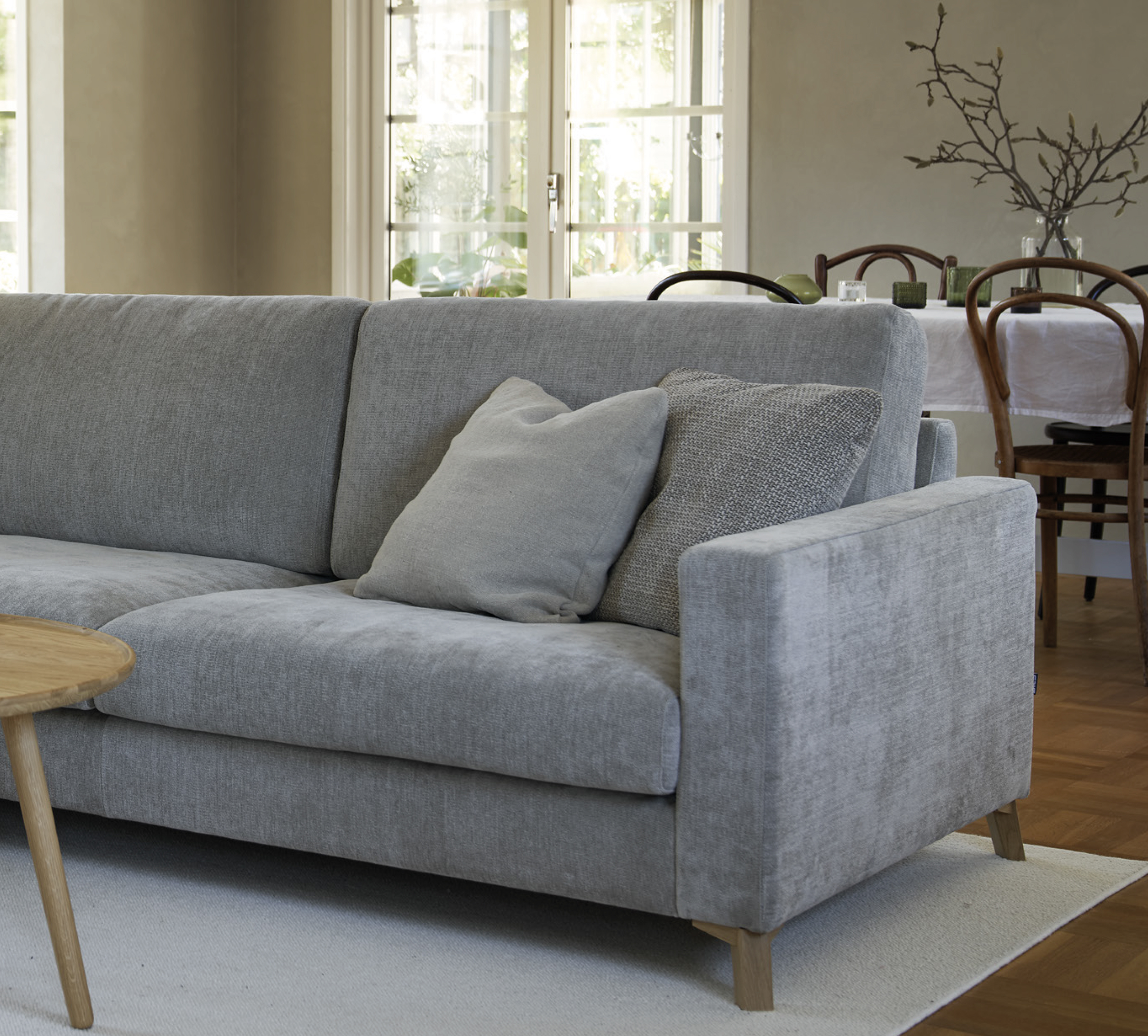 Canapé gris clair avec deux oreillers dans un salon, table basse en bois et salle à manger en arrière-plan. Canape Paris by Atelier Bis