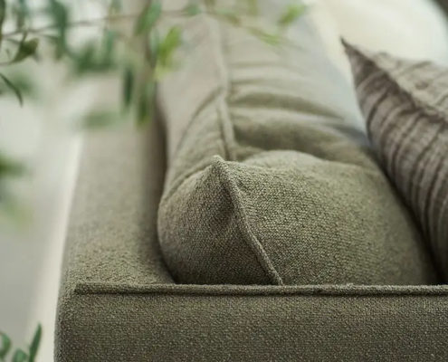 Gros plan d'un canapé vert texturé avec des coussins, partiellement encadré par une plante verte feuillue au premier plan. Canape Paris by Atelier Bis