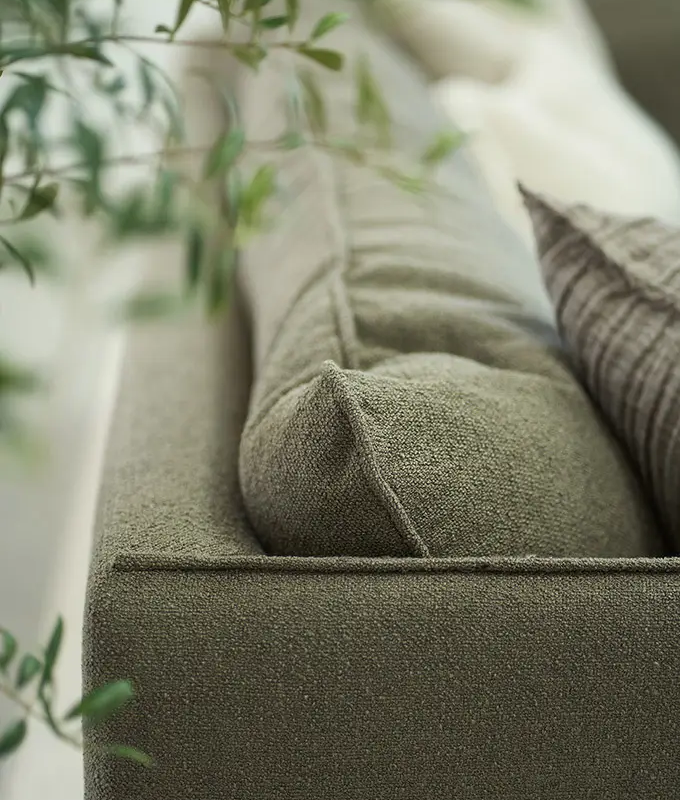 Gros plan d'un canapé vert texturé avec des coussins, partiellement encadré par une plante verte feuillue au premier plan. Canape Paris by Atelier Bis