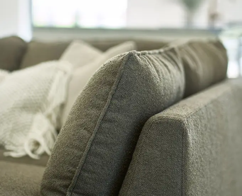 Gros plan sur un canapé gris avec une couverture blanche texturée drapée sur l'accoudoir, dans un salon lumineux. Canape Paris by Atelier Bis