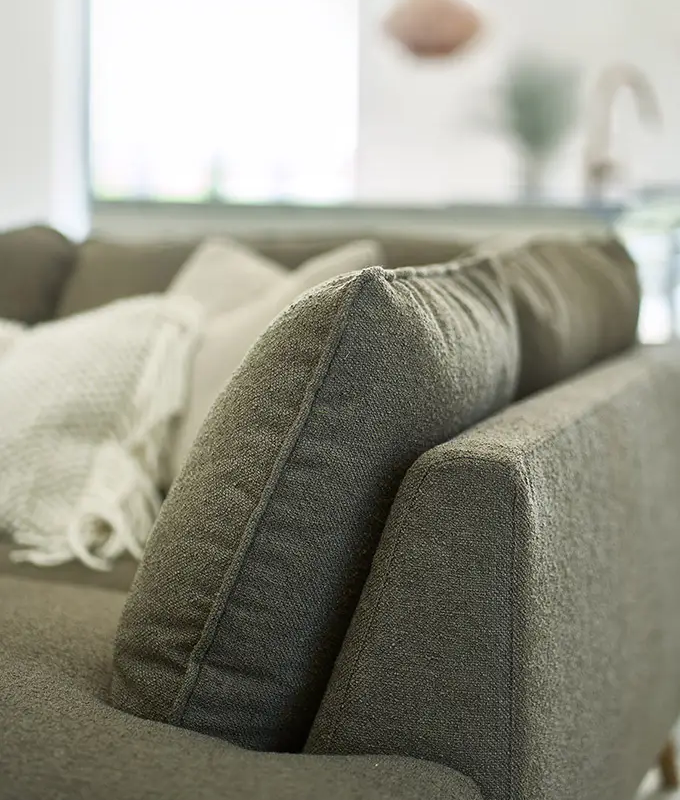 Gros plan sur un canapé gris avec une couverture blanche texturée drapée sur l'accoudoir, dans un salon lumineux. Canape Paris by Atelier Bis