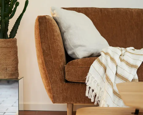 Un canapé marron avec un coussin blanc et une couverture rayée à côté d'un cactus dans un salon confortable. Canape Paris by Atelier Bis