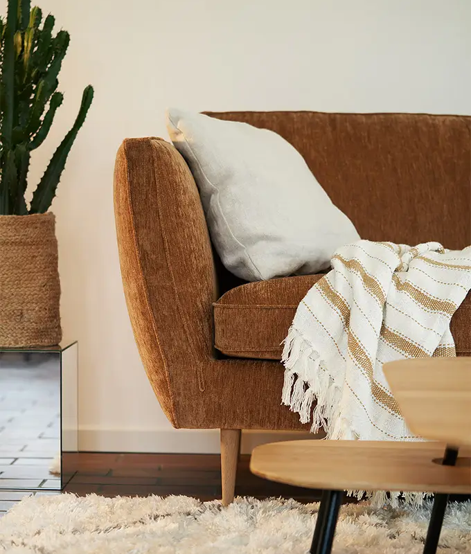 Un canapé marron avec un coussin blanc et une couverture rayée à côté d'un cactus dans un salon confortable. Canape Paris by Atelier Bis
