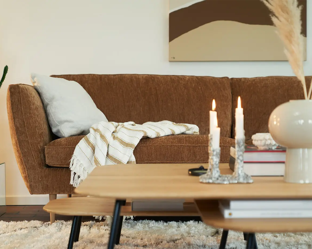 Canapé marron avec une couverture rayée, table basse avec des bougies et un cactus en pot dans un salon confortable. Canape Paris by Atelier Bis