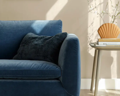 Salon moderne avec un canapé bleu, un coussin noir, une table d'appoint et des œuvres d'art murales sous la lumière naturelle du soleil. Canape Paris by Atelier Bis