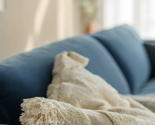 Une couverture beige à franges drapée sur l'accoudoir d'un canapé bleu dans un salon ensoleillé. Canape Paris by Atelier Bis