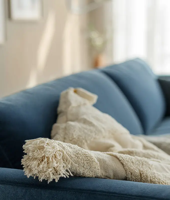 Une couverture beige à franges drapée sur l'accoudoir d'un canapé bleu dans un salon ensoleillé. Canape Paris by Atelier Bis