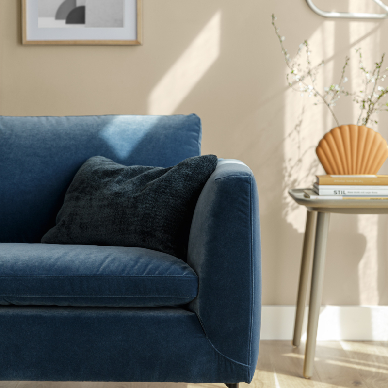 Canapé en velours bleu avec coussin foncé à côté d'une table d'appoint et d'un tableau mural dans un salon ensoleillé. Canape Paris by Atelier Bis