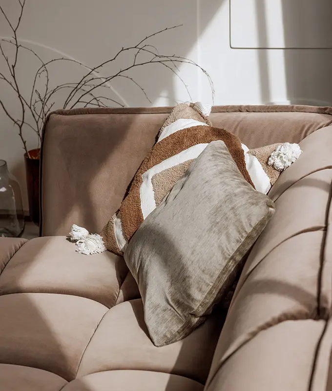 Canapé beige avec deux coussins décoratifs, l'un rayé et l'autre gris, au soleil avec des branches en arrière-plan. Canape Paris by Atelier Bis