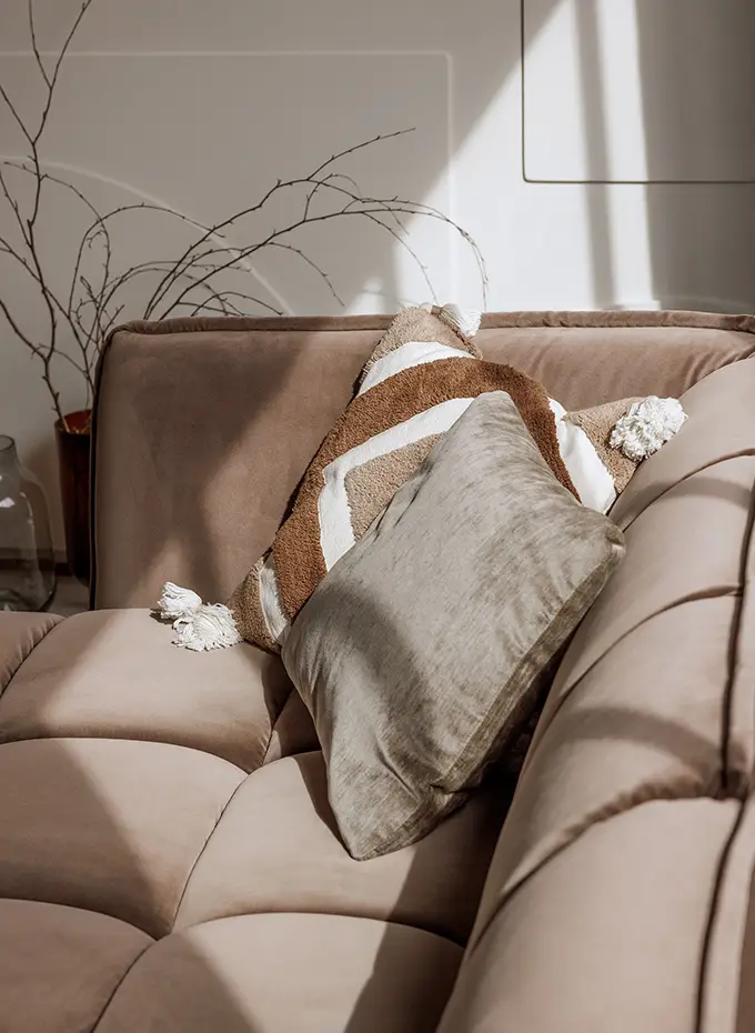 Canapé beige avec deux coussins décoratifs, l'un rayé et l'autre gris, au soleil avec des branches en arrière-plan. Canape Paris by Atelier Bis