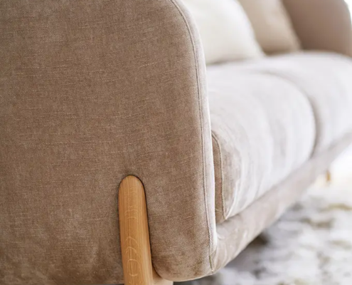 Gros plan sur un canapé beige avec des pieds en bois, posé sur un tapis blanc moelleux dans une pièce lumineuse. Canape Paris by Atelier Bis