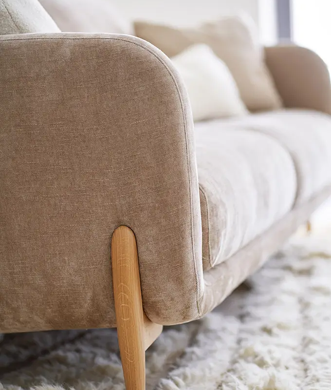 Gros plan sur un canapé beige avec des pieds en bois, posé sur un tapis blanc moelleux dans une pièce lumineuse. Canape Paris by Atelier Bis