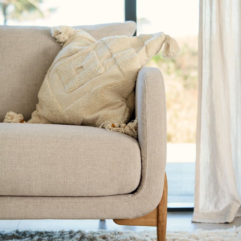 Canapé beige avec un coussin crème texturé près d'une fenêtre et d'un rideau léger sur un tapis moelleux. Canape Paris by Atelier Bis