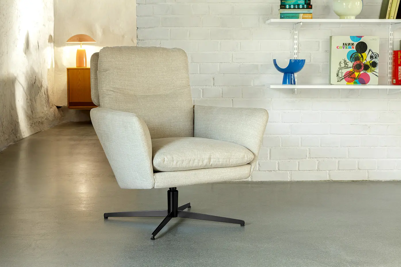Fauteuil pivotant rembourré beige sur base métallique dans une pièce moderne avec des étagères et une lampe. Canape Paris by Atelier Bis