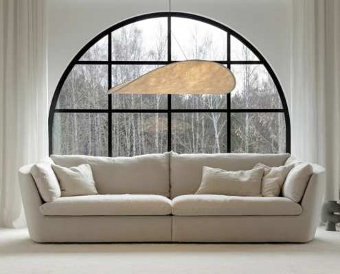 Un canapé beige moderne devant une grande fenêtre cintrée avec des arbres à l'extérieur et un décor minimaliste à l'intérieur. Canape Paris by Atelier Bis