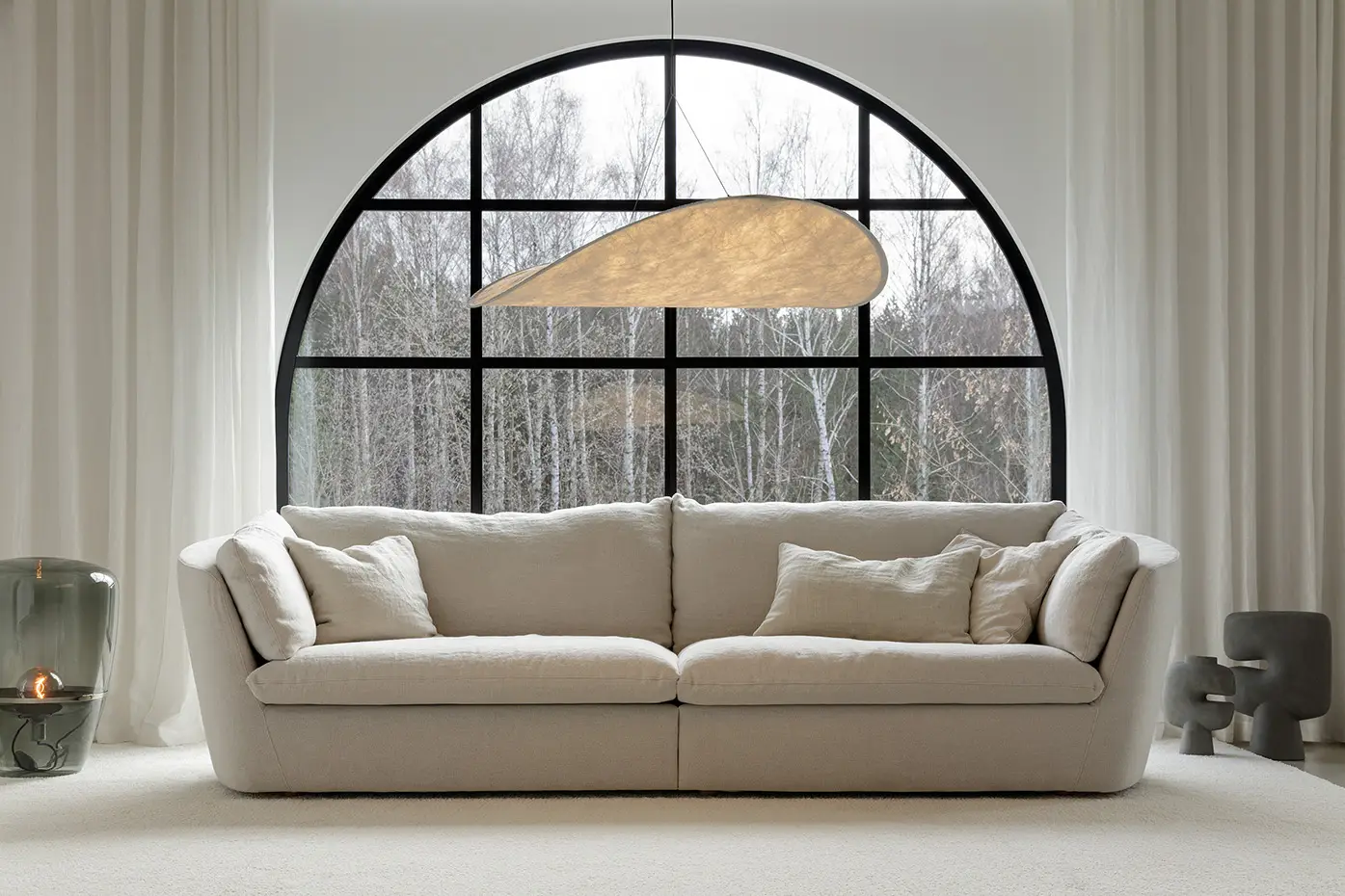 Un canapé beige moderne devant une grande fenêtre cintrée avec des arbres à l'extérieur et un décor minimaliste à l'intérieur. Canape Paris by Atelier Bis