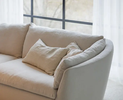 Canapé incurvé beige clair avec coussins devant une grande fenêtre avec des rideaux blancs transparents. Canape Paris by Atelier Bis