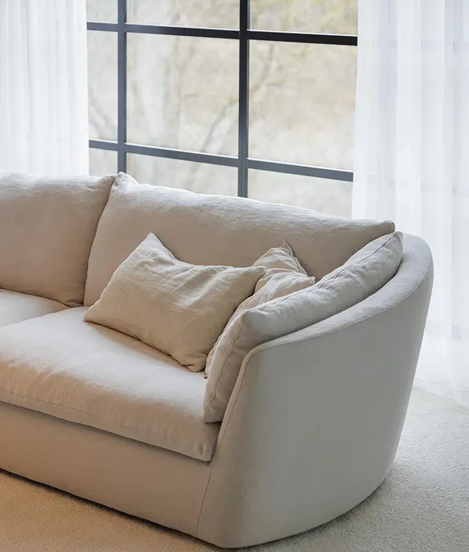Canapé incurvé beige clair avec coussins devant une grande fenêtre avec des rideaux blancs transparents. Canape Paris by Atelier Bis