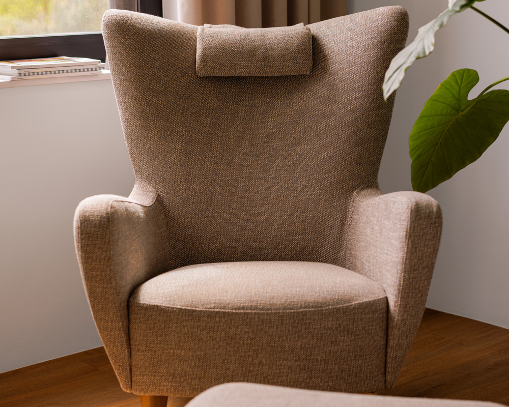 Un fauteuil beige avec appui-tête est placé près d'une fenêtre, à côté d'une grande plante verte. Canape Paris by Atelier Bis