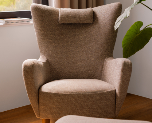 Un fauteuil beige avec appui-tête est placé près d'une fenêtre, à côté d'une grande plante verte. Canape Paris by Atelier Bis
