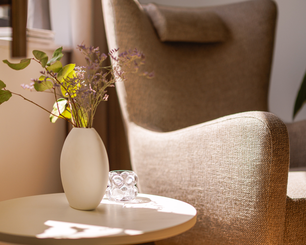 Fauteuil beige à côté d'une petite table avec un vase de fleurs blanches, dans une pièce confortable et ensoleillée. Canape Paris by Atelier Bis