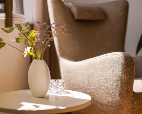 Fauteuil beige à côté d'une petite table avec un vase de fleurs blanches, dans une pièce confortable et ensoleillée. Canape Paris by Atelier Bis