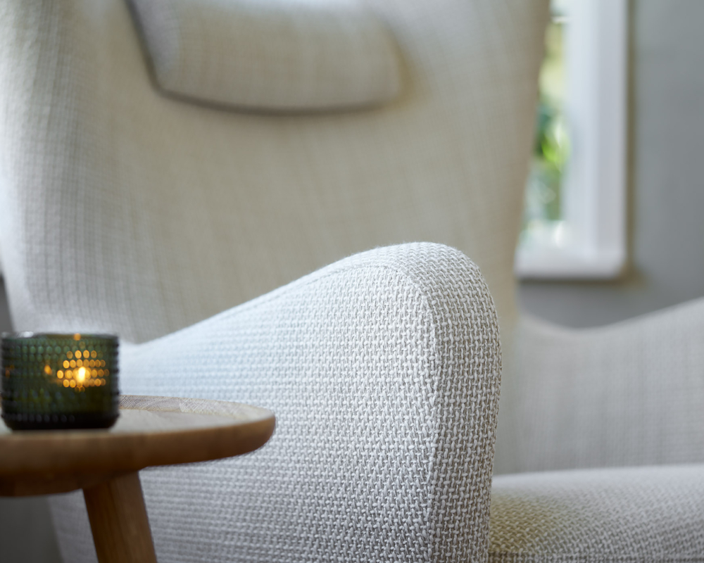 Gros plan d'un fauteuil gris clair texturé à côté d'une table en bois avec une bougie allumée dans un support en verre. Canape Paris by Atelier Bis
