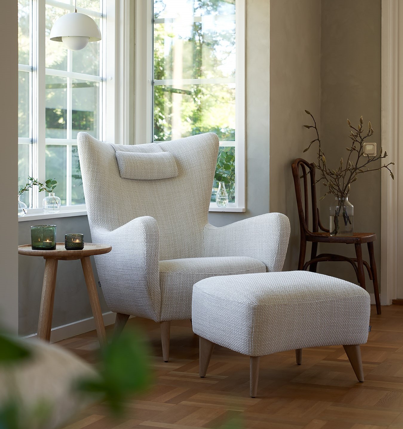 Fauteuil blanc confortable avec ottoman assorti près des fenêtres ensoleillées, à côté d'une petite table en bois avec des bougies. Canape Paris by Atelier Bis