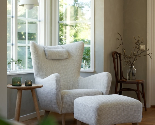 Un fauteuil blanc confortable avec un ottoman assorti près de grandes fenêtres et une petite table d'appoint en bois avec des bougies. Canape Paris by Atelier Bis