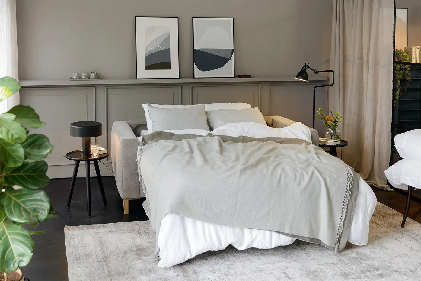 Une chambre à coucher confortable avec un lit moderne, une literie grise, des œuvres d'art murales, des plantes et un éclairage naturel doux. Canape Paris by Atelier Bis