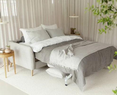 Canapé-lit moderne gris avec literie blanche et grise, tables d'appoint, plantes et lumière naturelle douce. Canape Paris by Atelier Bis