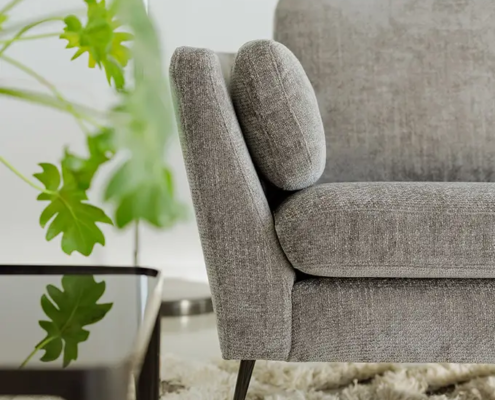 Gros plan sur un canapé gris moderne posé sur un tapis à poils longs, avec une plante verte à feuilles et une table noire au premier plan. Canape Paris by Atelier Bis