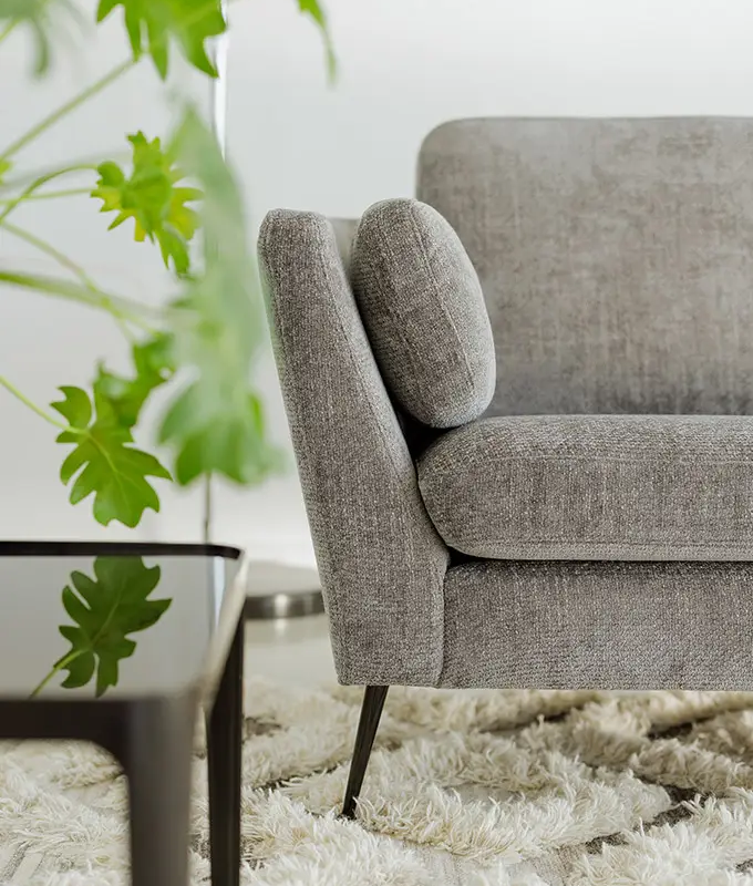 Gros plan sur un canapé gris moderne posé sur un tapis à poils longs, avec une plante verte à feuilles et une table noire au premier plan. Canape Paris by Atelier Bis