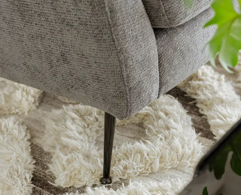 Gros plan d'un pied de canapé gris sur un tapis texturé à motifs crème et marron, avec des feuilles vertes au premier plan. Canape Paris by Atelier Bis