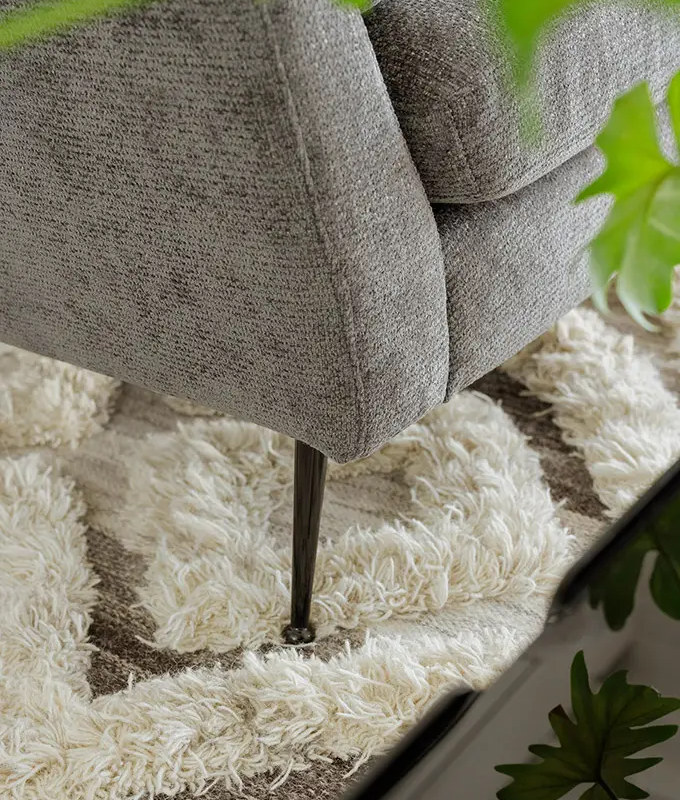 Gros plan d'un pied de canapé gris sur un tapis texturé à motifs crème et marron, avec des feuilles vertes au premier plan. Canape Paris by Atelier Bis